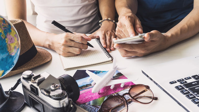 A couple sits at a desk with a camera, laptop, and a notepad as they plan a vacation
